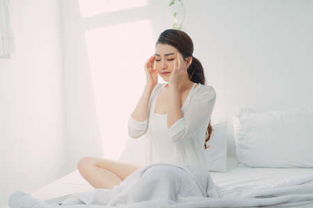 Young woman with headache, she is sitting on the bed and touching her templesの写真素材