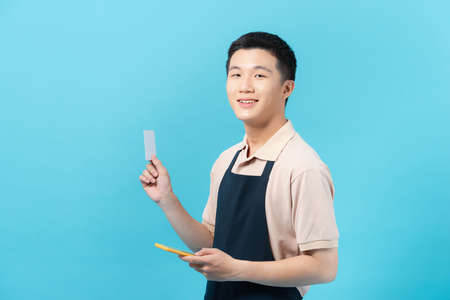 Cheerful young asian man wearing apron standing isolated over blue background, showing mobile phone and credit cardの写真素材