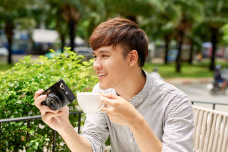 Smiling young male sitting at table in cafe with photo camera while drinking cup of coffeeの写真素材