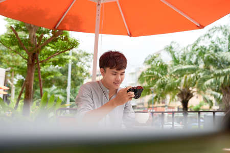 Smiling young male sitting at table in cafe with photo camera while drinking cup of coffeeの写真素材