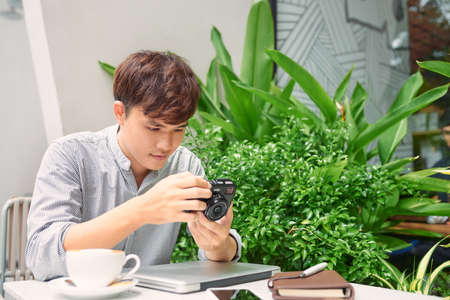 Successful hipster guy waiting friend in coffee shop while checking photos at vintage camera during free timeの写真素材