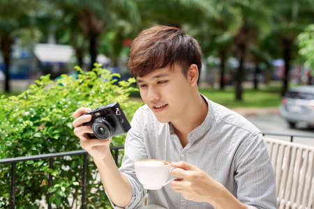Portrait of good-looking guy holding photo camera while sitting at the table with cup of coffeeの写真素材