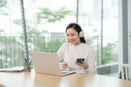 Smiling millennial female student in headphones sit at desk watch webinar making notesの写真素材