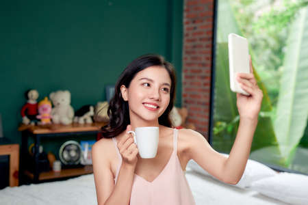 Beautiful young Asian woman using smartphone and holding a coffee cup while sitting on bed at home in the morningの写真素材
