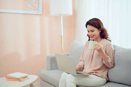 Morning concept. Excited female freelancer drinking coffee at home and using laptop, working online, free spaceの写真素材