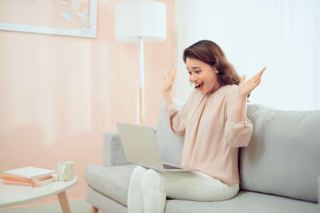 Happy smiling woman sitting at couch reading message looking at device screen using computer.の写真素材