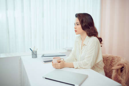 Asian female freelancer relaxing sitting with cup of coffee in front of laptopの写真素材
