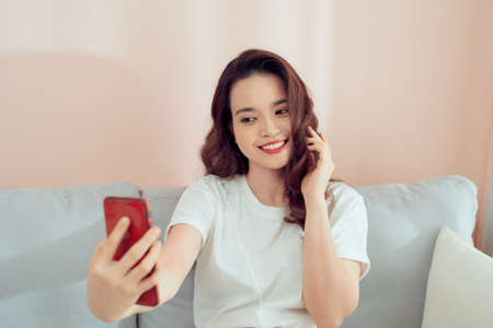 Shot of a happy young Asia woman taking selfie with her cellphone while sitting at living room.の写真素材