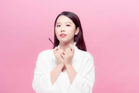 Dreaming asian woman with mascara for eyelashes with brush isolated over pink backgroundの写真素材