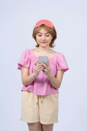 Dreamful young woman in pajamas sleep eye mask rest relaxing on mobile cell phone solated on white backgroundの写真素材