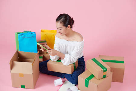 Young Asian woman business owner use your smartphone, get an order from customers, take notes, and make arrangements for delivery by post.の写真素材