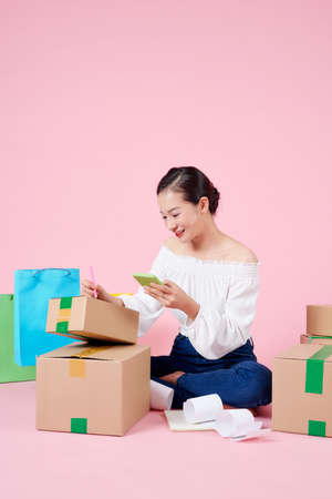 Smiling asian woman checking orders on smart phone and writing address on cardboard box.の写真素材