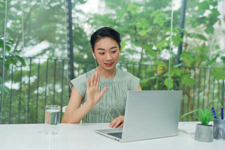 Head shot happy young  woman sitting at white desk, looking at camera, waving helloの写真素材