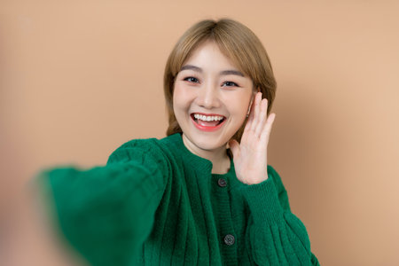 people concept - happy asian young woman taking selfie and waving hand over beige backgroundの写真素材