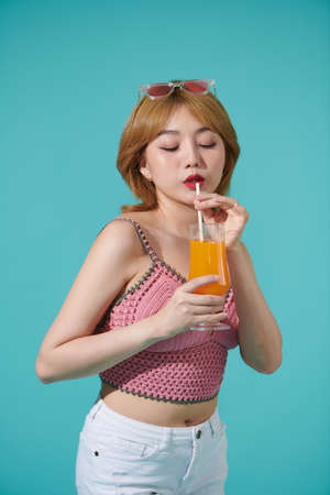 pretty young happy blonde woman wearing pink top and colourful sunglasses isolated over turquoise background holding orange juice cup drinking and enjoyingの写真素材