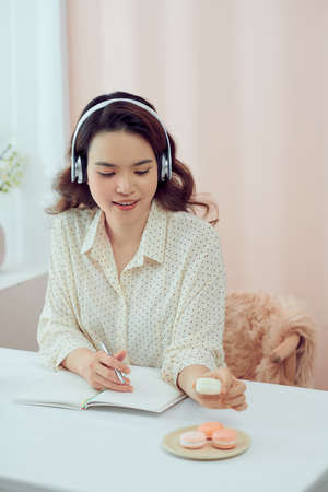 Asian young girl wearing headphones eating macarons and reading books at living roomの写真素材