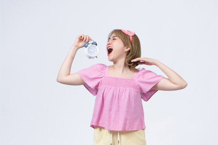 Tired and sleepy cute female in nightwear and sleep mask, holding red alarm clock, yawning turn away with closed eyesの写真素材