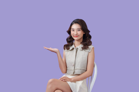 Young woman sitting on a chair over isolated violet background extending hands to the side for inviting to comeの写真素材