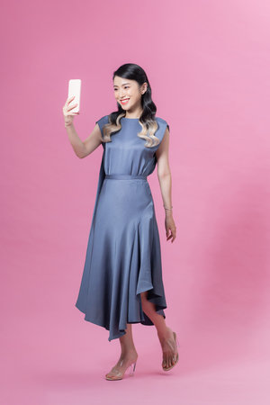 Beautiful happy young woman standing and taking selfie with smartphone over pink backgroundの写真素材