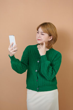 Asian woman hand holding smart phone wonder, thinking and planning something standing over beige backgroundの写真素材