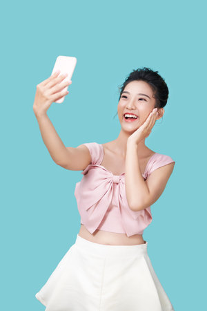 Happy young woman making silly faces while taking selfie on smartphone app with filters, standing against blue backgroundの写真素材