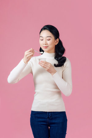 young beautiful woman eating yogurt with spoon looking satisfied healthy lifestyle isolated pink...