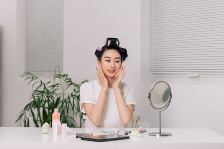 Pretty asian woman making face-lifting massage, sitting at table in bedroom, loking at mirror and smilingの写真素材