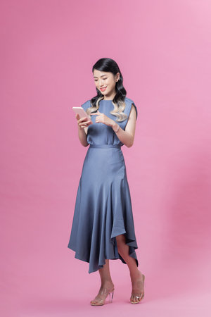Full length young asian woman with hold mobile cell phone chat online walk go isolated on pink backgroundの写真素材