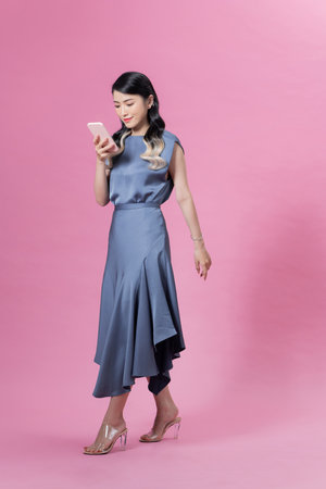 Full body side portrait of relaxed young woman walking and reading text message on the mobile phone over pink backgroundの写真素材