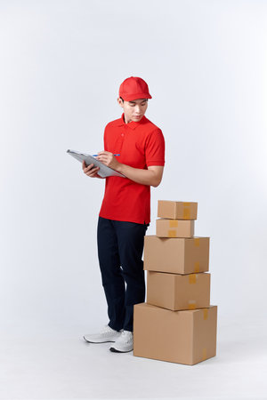 Cheerful professional male mover in casual wear making notes, holding clipboard, standing near a stack of cardboard boxes while doing home relocation. Service conceptの写真素材