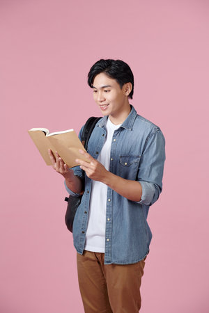 A young student carrying a backpack and holding an open book reading, isolated on a pink background.の写真素材
