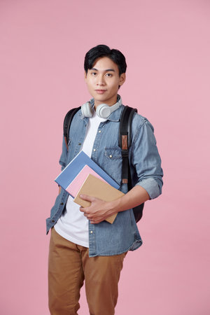 Positive young man with backpack, headphones and notebooks posing on pink backgroundの写真素材