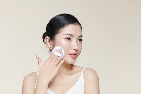 Beautiful young woman applying face powder with puff applicator over beige backgroundの写真素材