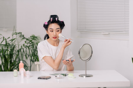 Beautiful woman makes blush on her face using makeup brush while sitting at home in a room at a table and looking in the mirrorの写真素材