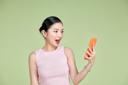 Portrait of surprised young Asian woman using mobile phone on green backgroundの写真素材