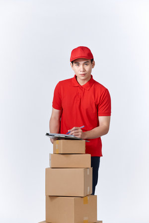 Cheerful professional male mover in casual wear making notes, holding clipboard, standing near a stack of cardboard boxes while doing home relocation. Service conceptの写真素材