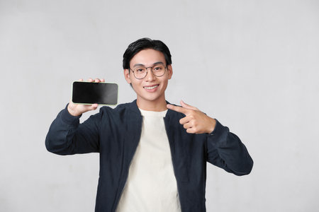 Smiling young man holds a modern smartphone in his hand and shows his finger on a blank screen,の写真素材