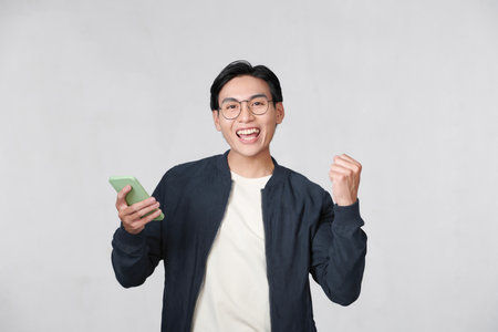 Excited young Asian man using smartphone and making winner gesture isolatedの写真素材
