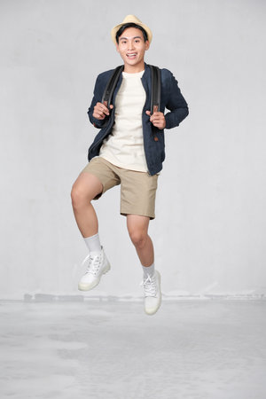 Happy smiling young Asian tourist man with backpack jumping in mid-airの写真素材