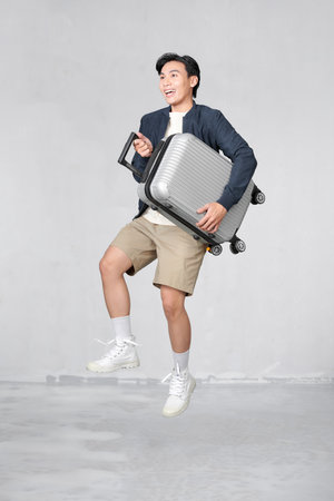 Jumping portrait of young fit healthy smiling handsome Asian tourist man with baggage ready to flyの写真素材
