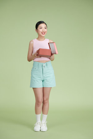 Portrait of a cheerful asian woman opening gift box over green backgroundの写真素材