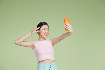 Image of pleased asian woman laughing and showing peace sign while taking selfie photo on cellphoneの写真素材