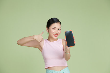 Portrait of smiling asian woman holding smartphone with black blank device screen in hand, showing thumb up gestureの写真素材