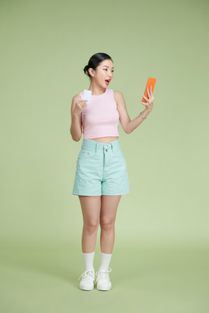 Happy young girl showing plastic credit card while holding mobile phone isolated over green backgroundの写真素材
