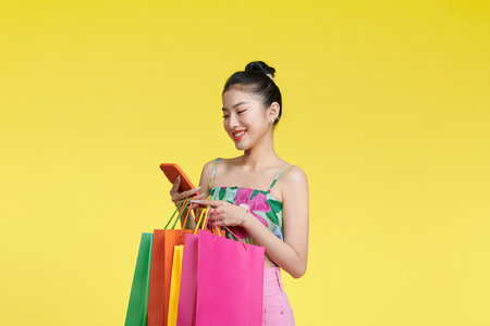 Portrait of a cheerful young girl in summer clothes holding mobile phone with shopping bags isolated over yellow backgroundの写真素材