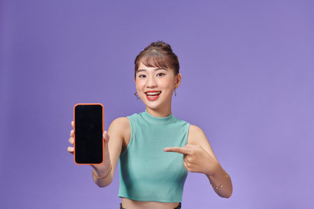 Young Asian woman holding phone in hand and pointing at it with playful expressionの写真素材