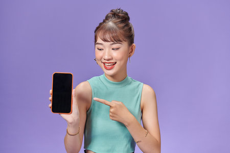 Cheerful cute young woman holding and pointing on blank screen mobile phoneの写真素材