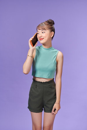 Young girl calling on mobile phone while standing and looking away isolated over purple backgroundの写真素材