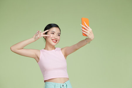 Smiling adorable Asian female making selfie photo on smartphone with positive expression over green backgroundの写真素材