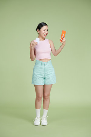Happy young girl showing plastic credit card while holding mobile phone isolated over green backgroundの写真素材